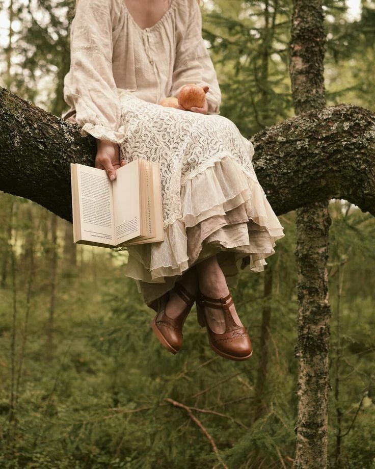 Person in a cream dress reading a book while seated on a tree branch in a forest